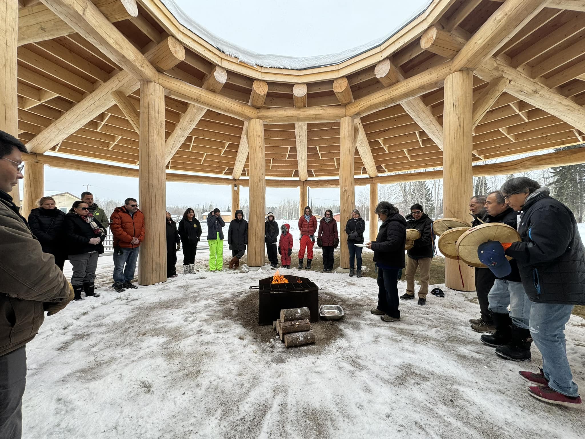 Adults and children dressed in snow gear gather around a small fire inside a wooden structure. The structure is made of two circles of external and internal poles. Beams, extending from the external poles to the internal, support an angled roof with a large central opening. The people stand in a wide circle underneath the roof while the fire burns below the opening. Snow dusts the ground and four people play hand drums.